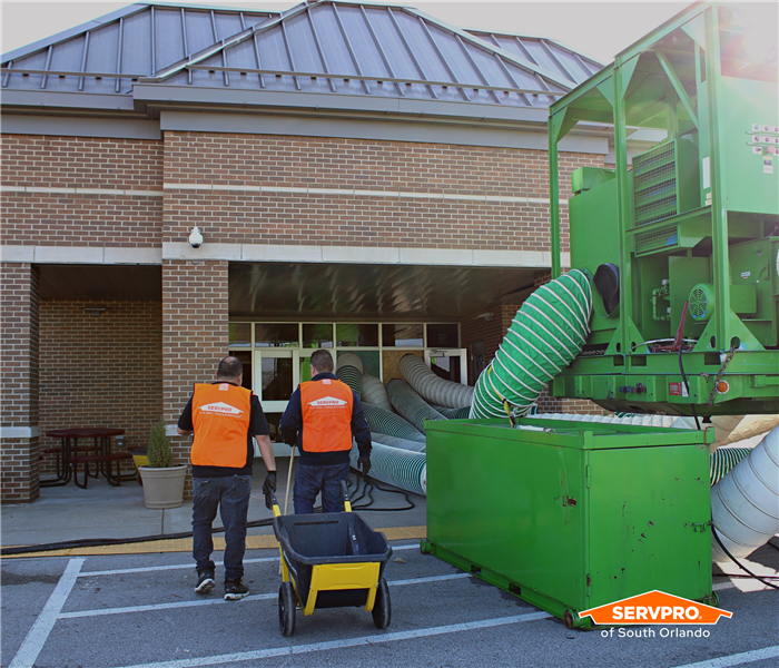 Two SERVPRO of South Orlando crew in orange vests walking into a school next to a big, green desiccant dehumidifier 